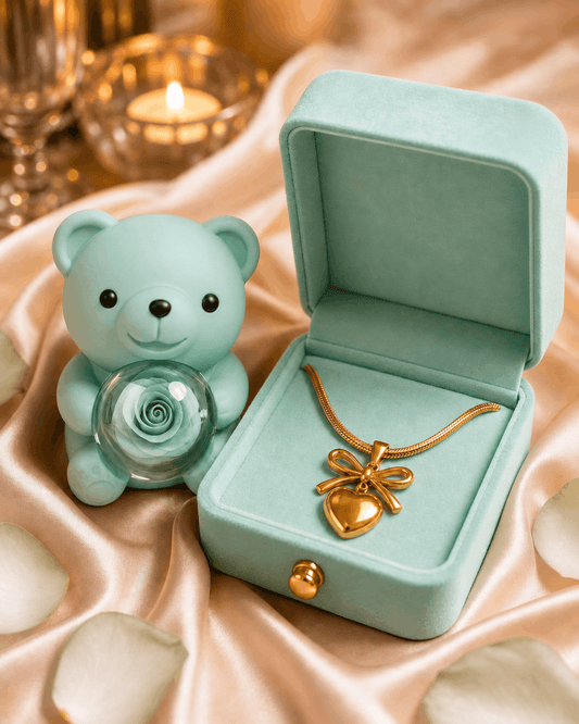 Open teal jewelry box with a gold necklace, next to a small green teddy bear with a glass rose on a pink fabric background.